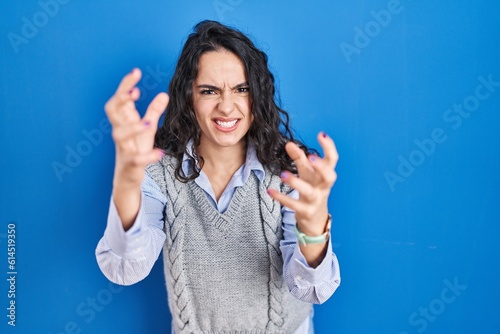 Young brunette woman standing over blue background shouting frustrated with rage, hands trying to strangle, yelling mad