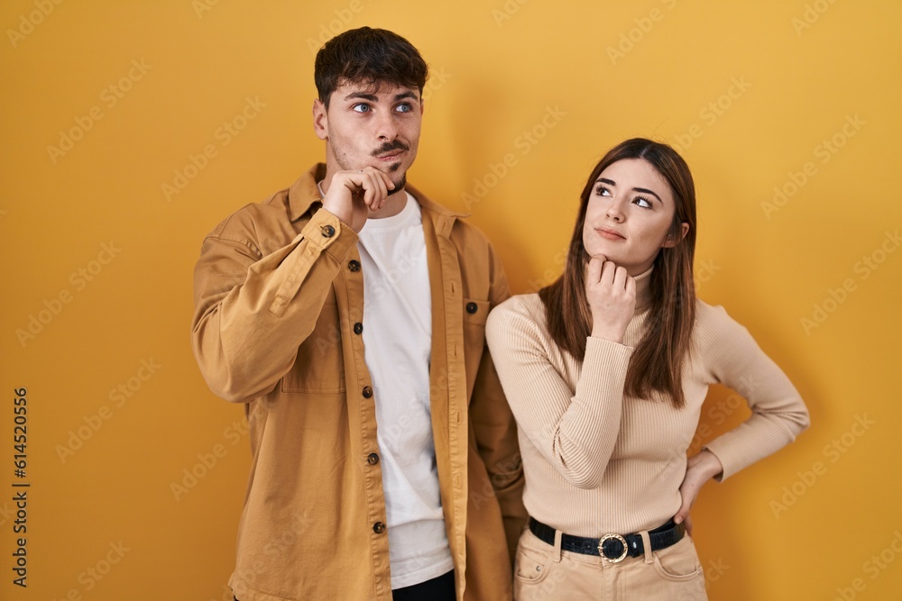 Young hispanic couple standing over yellow background with hand on chin thinking about question, pensive expression. smiling with thoughtful face. doubt concept. Young hispanic couple standing over yellow background with hand on chin thinking about question, pensive expression. smiling with thoughtful face. doubt concept.