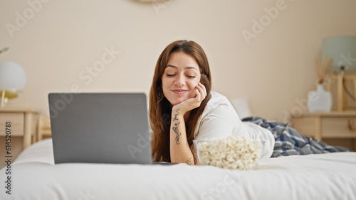 Young beautiful hispanic woman using laptop eating popcorn lying on bed at bedroom