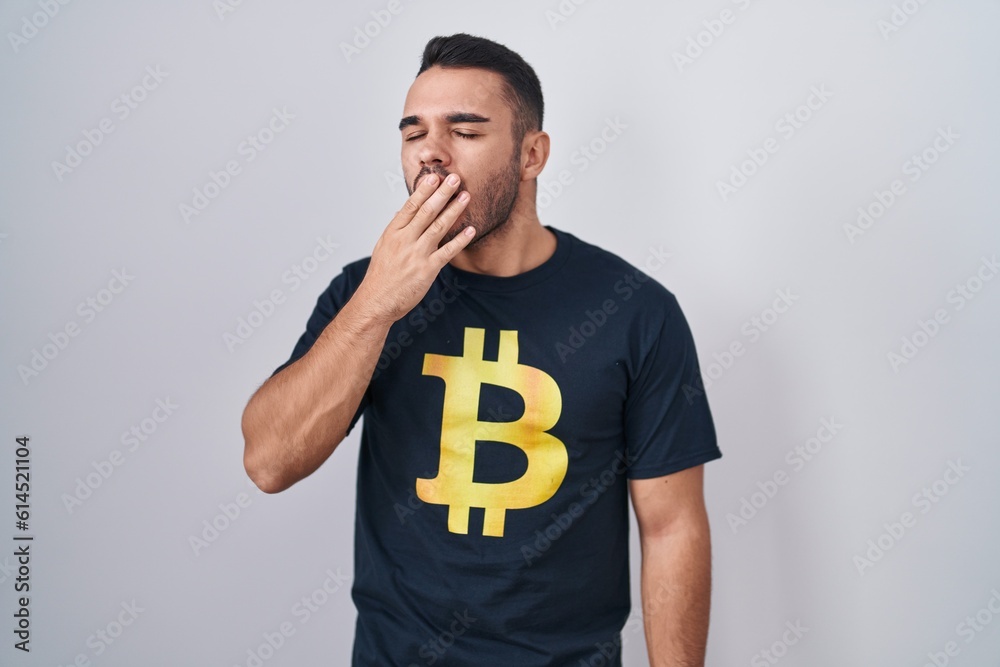 Young hispanic man wearing bitcoin t shirt bored yawning tired covering mouth with hand. restless and sleepiness. Young hispanic man wearing bitcoin t shirt bored yawning tired covering mouth with hand. restless and sleepiness.