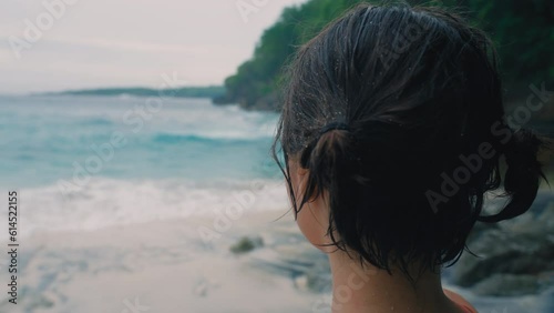 Happy Korean girl after swimming in sea points her finger at swirling waves looks into camera and pointedly nods her head. A delightful woman against background of crashing ocean waves on wild beach. photo