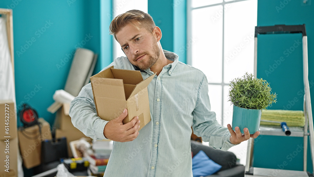 Young caucasian man holding package and plant talking on smartphone at new home