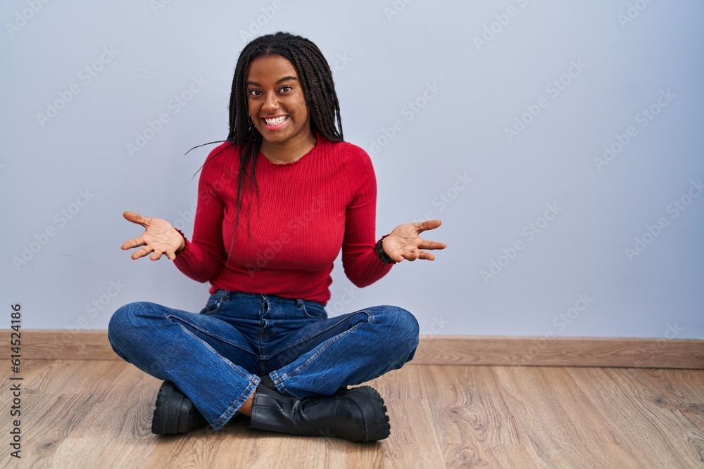 Young african american with braids sitting on the floor at home smiling cheerful with open arms as friendly welcome, positive and confident greetings Young african american with braids sitting on the floor at home smiling cheerful with open arms as friendly welcome, positive and confident greetings