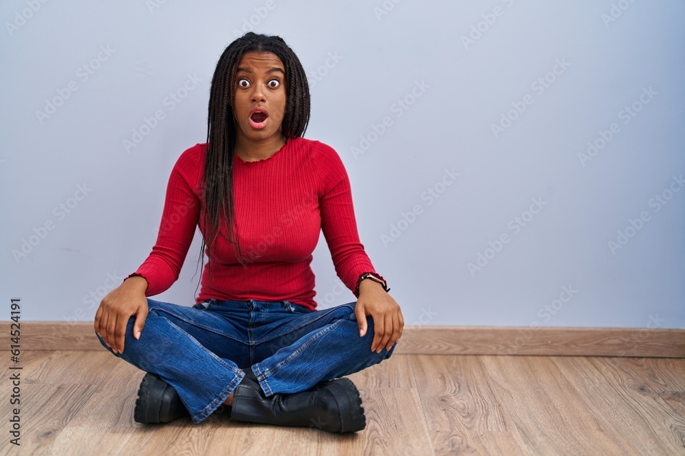 Young african american with braids sitting on the floor at home in shock face, looking skeptical and sarcastic, surprised with open mouth Young african american with braids sitting on the floor at home in shock face, looking skeptical and sarcastic, surprised with open mouth