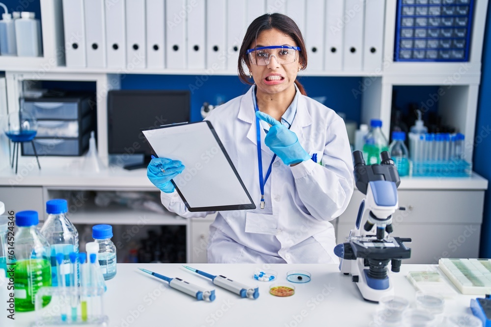 Hispanic young woman working at scientist laboratory pointing aside worried and nervous with forefinger, concerned and surprised expression