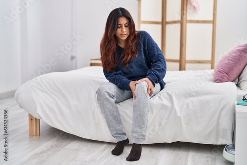 Young hispanic woman suffering for knee pain sitting on bed at bedroom
