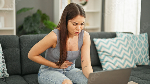 Young beautiful hispanic woman suffering for stomach ache using laptop at home