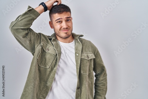 Young hispanic man standing over isolated background confuse and wondering about question. uncertain with doubt, thinking with hand on head. pensive concept.