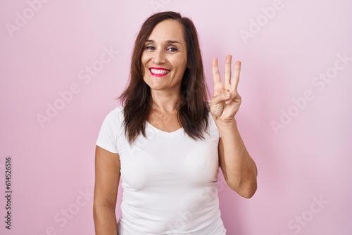 Middle age brunette woman standing over pink background showing and pointing up with fingers number three while smiling confident and happy.