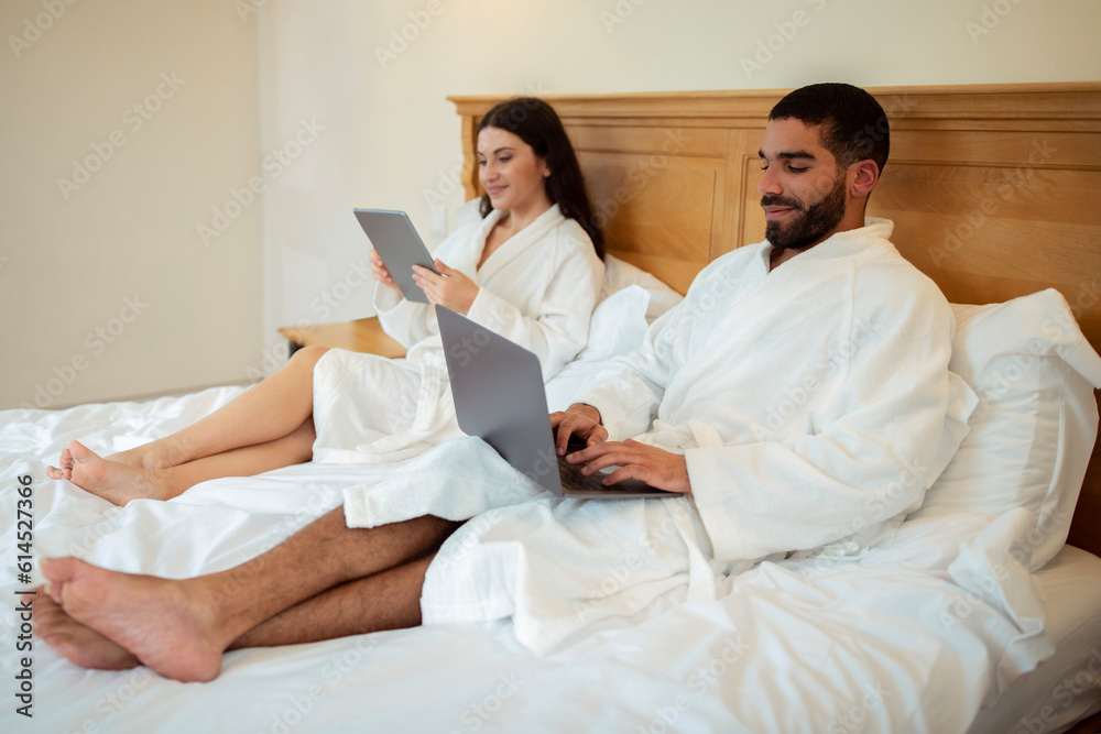 Husband And Wife Browsing Internet On Computers Lying In Bedroom
