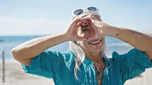 Middle age grey-haired woman tourist smiling confident doing heart gesture with hands at beach