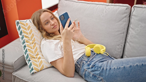 Young blonde woman using smartphone lying on the sofa at home