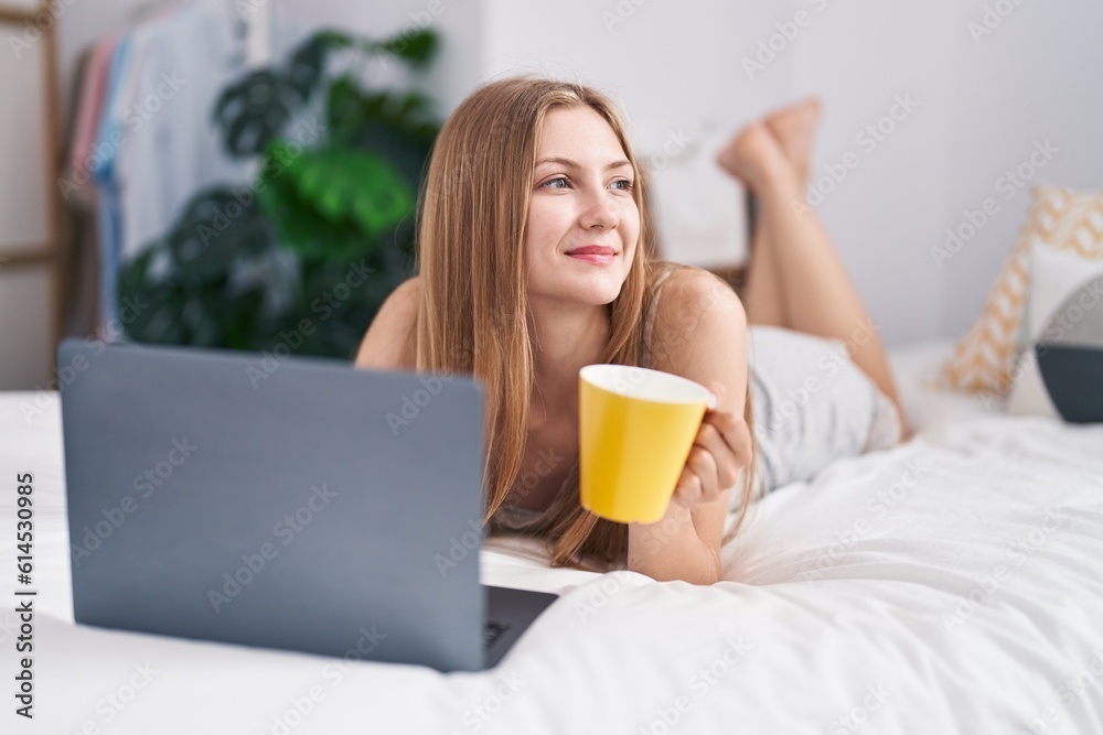 Young caucasian woman using laptop drinking coffee lying on bed at bedroom