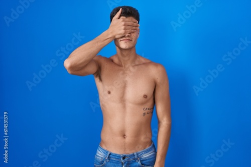 Young hispanic man standing shirtless over blue background covering eyes with hand, looking serious and sad. sightless, hiding and rejection concept