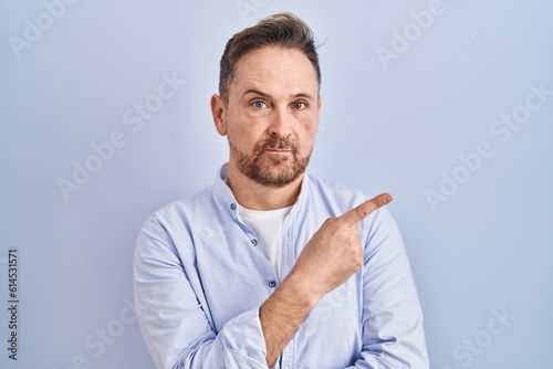 Middle age caucasian man standing over blue background pointing with hand finger to the side showing advertisement  serious and calm face