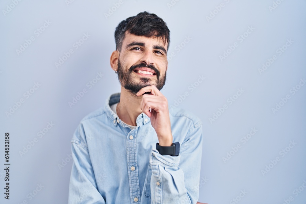 Young hispanic man with beard standing over blue background with hand on chin thinking about question, pensive expression. smiling and thoughtful face. doubt concept. Young hispanic man with beard standing over blue background with hand on chin thinking about question, pensive expression. smiling and thoughtful face. doubt concept.