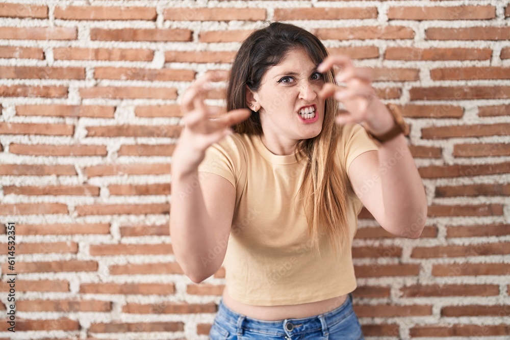 Young brunette woman standing over bricks wall shouting frustrated with rage, hands trying to strangle, yelling mad