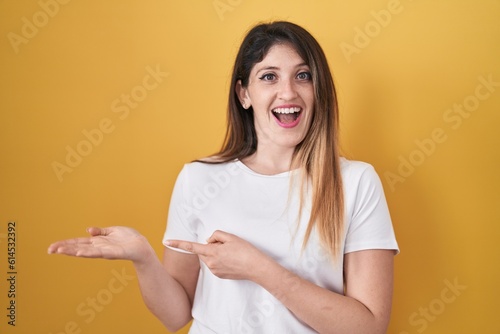 Young brunette woman standing over yellow background amazed and smiling to the camera while presenting with hand and pointing with finger.