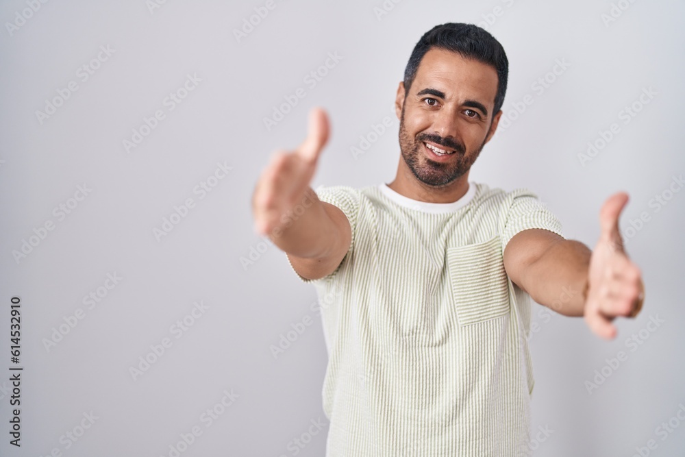 Hispanic man with beard standing over isolated background looking at the camera smiling with open arms for hug. cheerful expression embracing happiness. Hispanic man with beard standing over isolated background looking at the camera smiling with open arms for hug. cheerful expression embracing happiness.