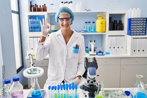 Brunette woman working at scientist laboratory smiling positive doing ok sign with hand and fingers. successful expression.