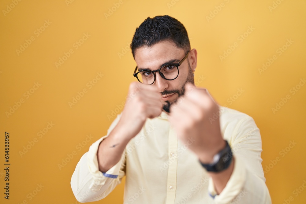 Hispanic young man wearing business clothes and glasses ready to fight with fist defense gesture, angry and upset face, afraid of problem Hispanic young man wearing business clothes and glasses ready to fight with fist defense gesture, angry and upset face, afraid of problem