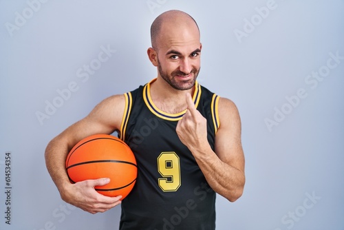Young bald man with beard wearing basketball uniform holding ball beckoning come here gesture with hand inviting welcoming happy and smiling