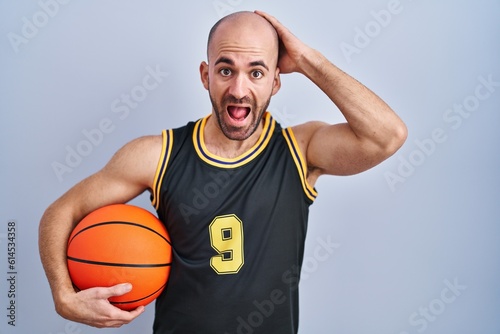 Young bald man with beard wearing basketball uniform holding ball crazy and scared with hands on head, afraid and surprised of shock with open mouth