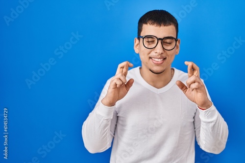 Young arab man wearing casual white shirt and glasses gesturing finger crossed smiling with hope and eyes closed. luck and superstitious concept.