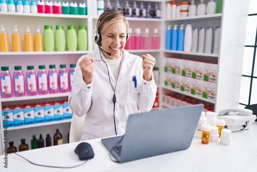 Young caucasian woman working at pharmacy drugstore using laptop very happy and excited doing winner gesture with arms raised, smiling and screaming for success. celebration concept.
