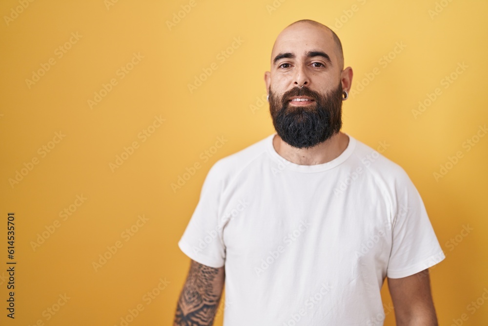 Young hispanic man with beard and tattoos standing over yellow background relaxed with serious expression on face. simple and natural looking at the camera. Young hispanic man with beard and tattoos standing over yellow background relaxed with serious expression on face. simple and natural looking at the camera.