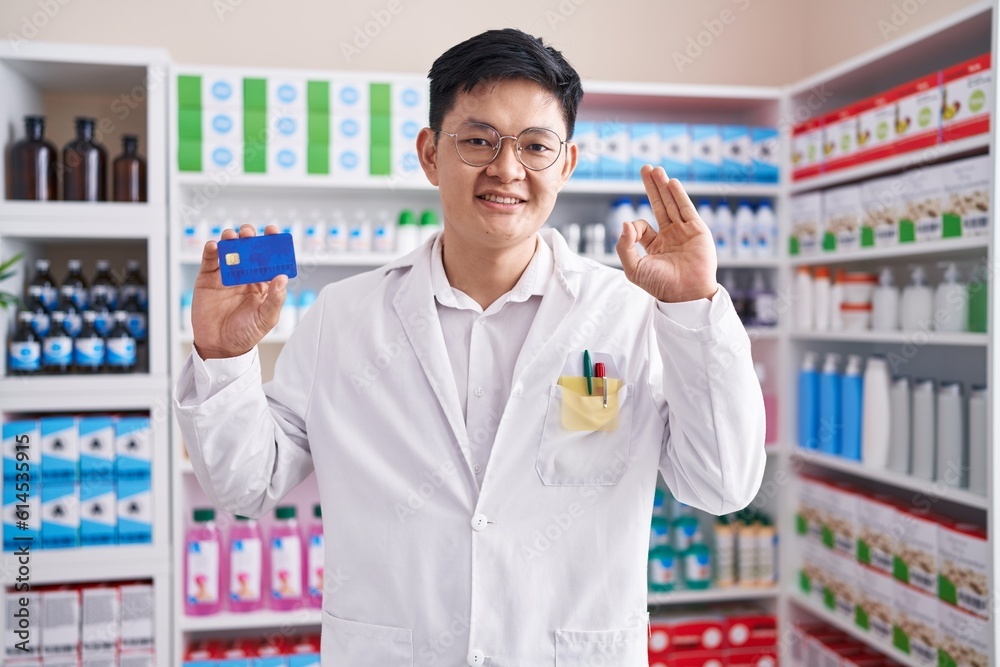 Young asian man working at pharmacy drugstore holding credit card doing ok sign with fingers, smiling friendly gesturing excellent symbol