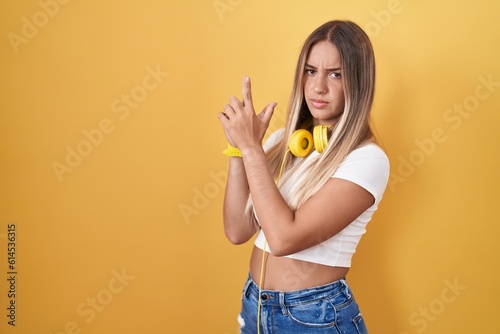 Young blonde woman standing over yellow background wearing headphones holding symbolic gun with hand gesture, playing killing shooting weapons, angry face