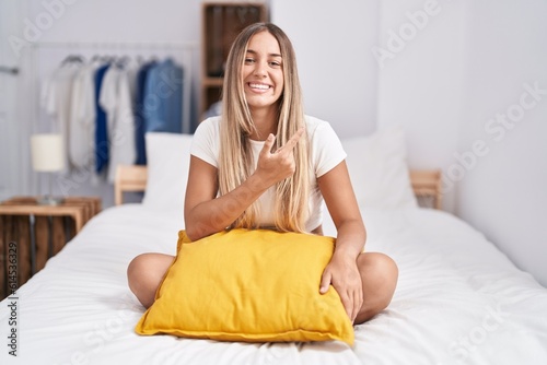 Young blonde woman sitting on the bed with pillow at home cheerful with a smile of face pointing with hand and finger up to the side with happy and natural expression on face