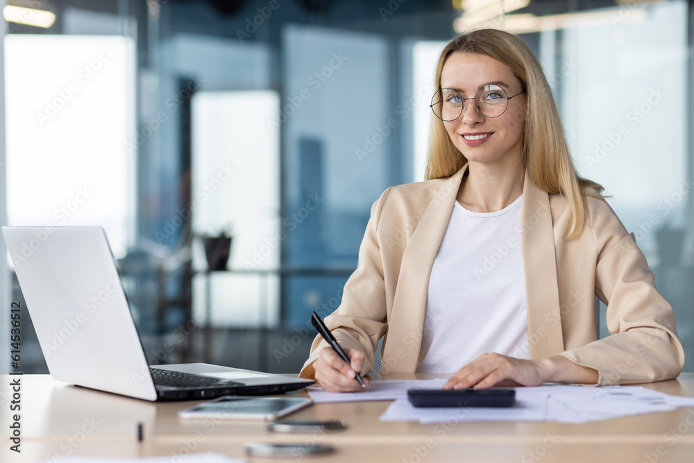 Portrait of a young female student, intern. Sitting in the office at the table, studying online from a laptop, looking at the camera with a smile Portrait of a young female student, intern. Sitting in the office at the table, studying online from a laptop, looking at the camera with a smile