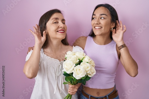 Hispanic mother and daughter holding bouquet of white flowers smiling with hand over ear listening an hearing to rumor or gossip. deafness concept.