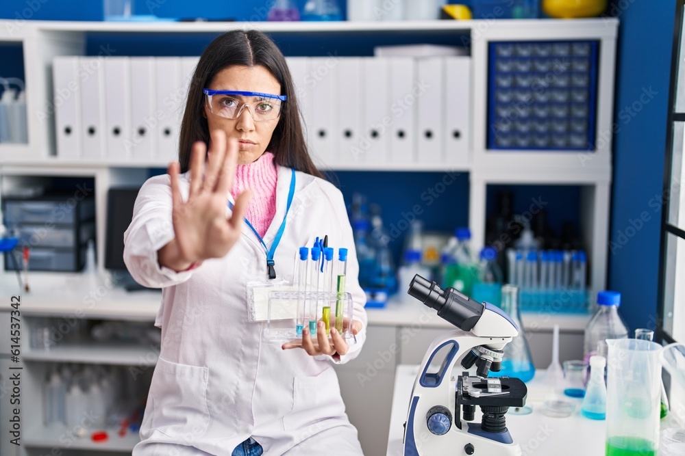 Young brunette woman working at scientist laboratory with open hand doing stop sign with serious and confident expression, defense gesture