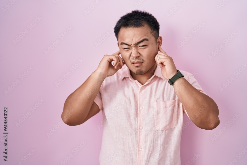 Chinese young man standing over pink background covering ears with fingers with annoyed expression for the noise of loud music. deaf concept. Chinese young man standing over pink background covering ears with fingers with annoyed expression for the noise of loud music. deaf concept.