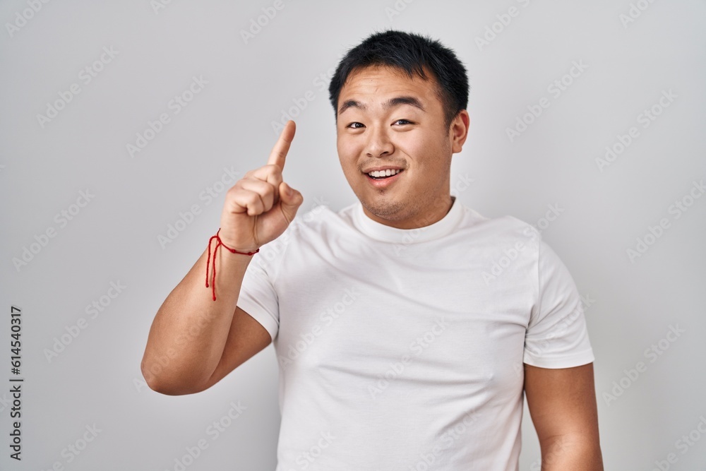 Young chinese man standing over white background pointing finger up with successful idea. exited and happy. number one. Young chinese man standing over white background pointing finger up with successful idea. exited and happy. number one.