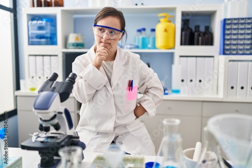 Hispanic girl with down syndrome working at scientist laboratory with hand on chin thinking about question, pensive expression. smiling with thoughtful face. doubt concept.