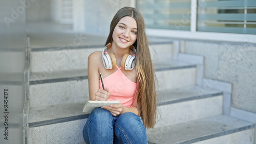 Young beautiful girl student taking notes sitting on stairs smiling at school
