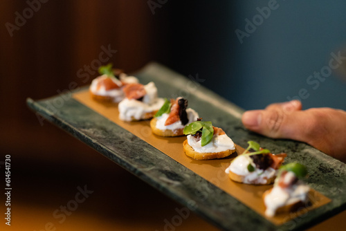 horizontal shot of server's hand holding dark serving tray of crostini with ricotta prosciutto and basil