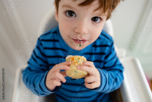 cute caucasian toddler boy in blue and black striped shirt sitting in white highchair holding a vanilla cupcake with crumbs on his face
