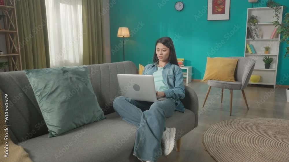 An attractive Asian woman sits on a sofa in the living room with a laptop on her lap. A woman is working, typing a text, a message, chatting. Remote work, freelancer. HDR BT2020 HLG Material. An attractive Asian woman sits on a sofa in the living room with a laptop on her lap. A woman is working, typing a text, a message, chatting. Remote work, freelancer. HDR BT2020 HLG Material.