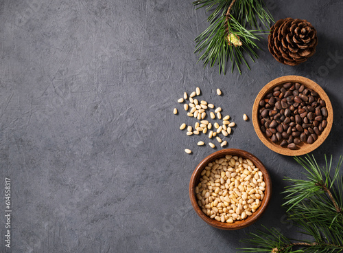 Pine nuts in a wooden bowl and scattered on a dark background with branches of pine needles and a cone. The concept of a natural, organic and healthy superfood and snack.