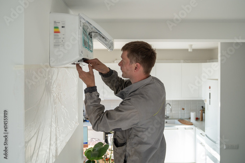 Young man technician worker in uniform fixing repairing apartment air conditioner, installing wall-mounted mini split. AC unit maintenance concept