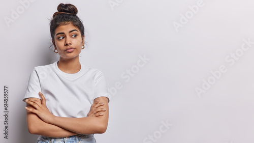 Studio shot of pleasant looking Iranian woman keeps arms folded concentrated aside with thoughtful expression wears casual t shirt isolated over white background copy space for your advertisement