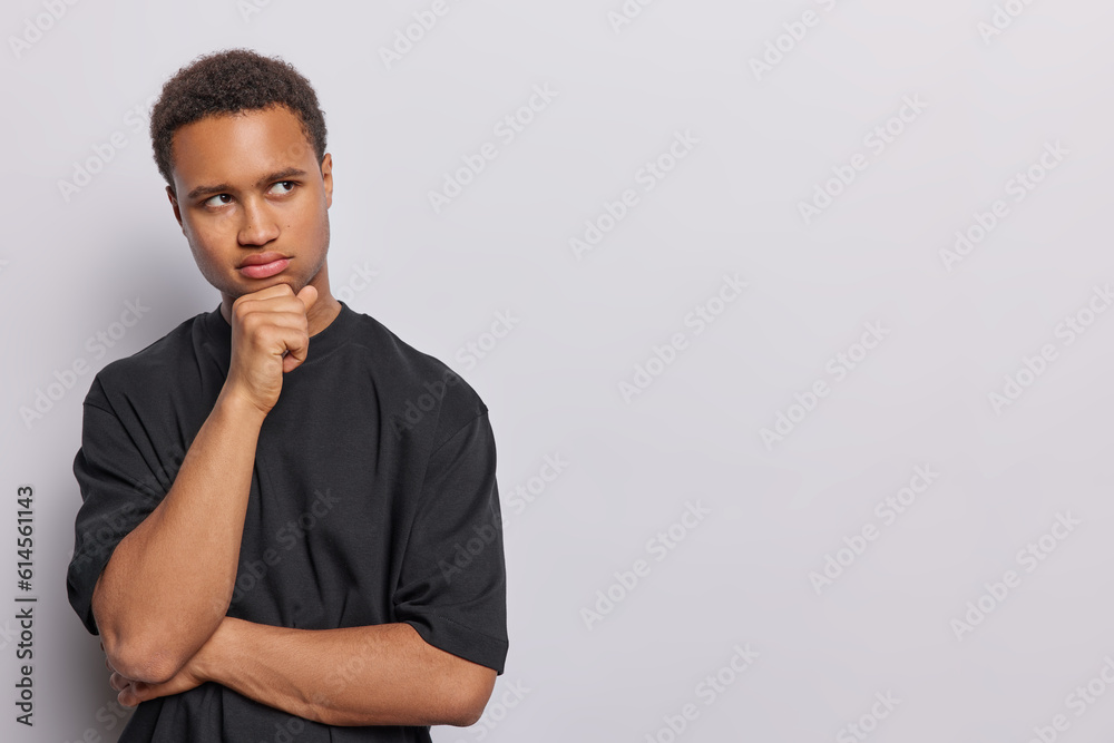 Horizontal shot of serious short haired man keeps hand on chin concentrated aside pensively has focused gaze at something wears black t shirt isolated over white background copy space aside. Horizontal shot of serious short haired man keeps hand on chin concentrated aside pensively has focused gaze at something wears black t shirt isolated over white background copy space aside.
