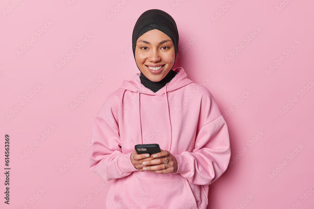 Pleased young Muslim woman using phone application edit pics with funny filters smiles positively wears black hijab and sweatshirt isolated over pink background. People and technology concept