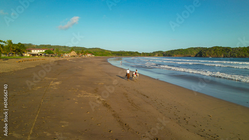 AERIAL: Scenic sandy beach with a family on a guided horseback riding adventure