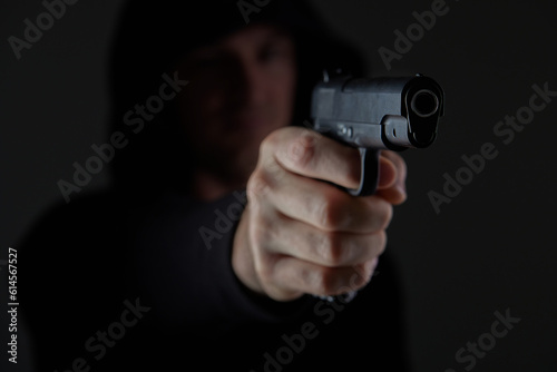 robber with gun aiming into camera. Man in hood threatens with firearm. Weapon in person's hands. Murderer or armed thief. Criminal with pistol.